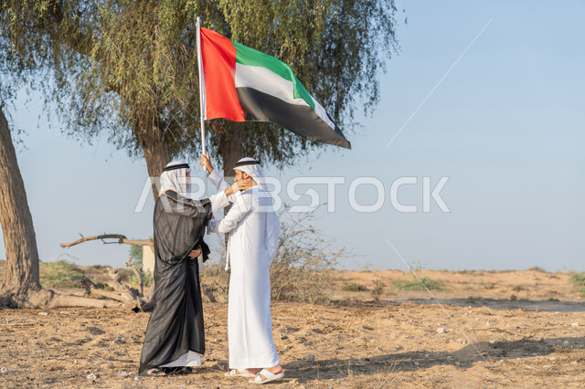 ارتداء الازياء الشعبية التقليدية للاحتفال بذكرى تأسيس الاتحاد 2 ديسمبر، يوم العلم 3 نوفمبر، حب الوطن والاعتزاز به، اليوم الوطني الاماراتي 1971، رجلان عربيان خليجيان اماراتيان يرتديان الزي التقليدي يرفعان علم الامارات العربية المتحدة في الصحراء، خلفية الطبيعة