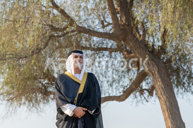 ذكرى تأسيس الاتحاد 2 ديسمبر، حب الوطن والاعتزاز به، استذكار اليوم الوطني الاماراتي 1971، رجل عربي خليجي إماراتي يرتدي الكندورة والبشت مع الغترة البيضاء يقف امام شجرة خضراء كبيرة في النهار، التجول في المناطق الطبيعية في الامارات العربية المتحدة، خلفية الطبيعة