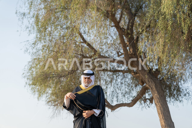 ذكرى تأسيس الاتحاد 2 ديسمبر، حب الوطن والاعتزاز به، استذكار اليوم الوطني الاماراتي 1971، رجل عربي خليجي إماراتي يرتدي الكندورة والبشت مع الغترة البيضاء يقف امام شجرة خضراء كبيرة في النهار، التجول في المناطق الطبيعية في الامارات العربية المتحدة، خلفية الطبيعة
