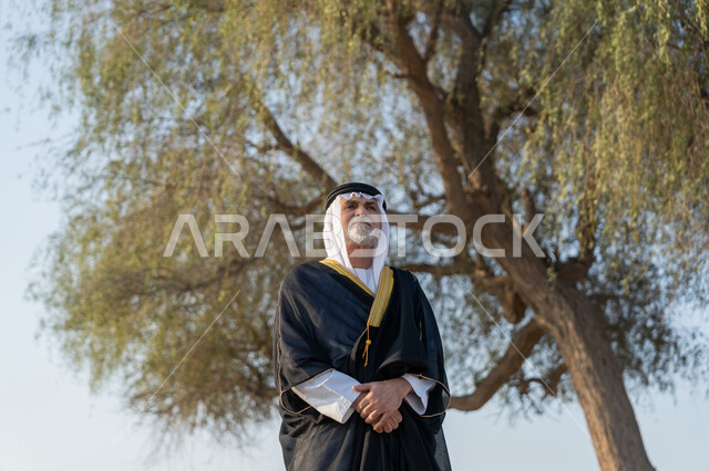Union Day December 2, love and pride in the homeland, remembering the UAE National Day 1971, an Arab Gulf Emirati man wearing a kandura and a bisht with a white ghutra standing in front of a large green tree during the day, walking in natural areas in the United Arab Emirates, nature background