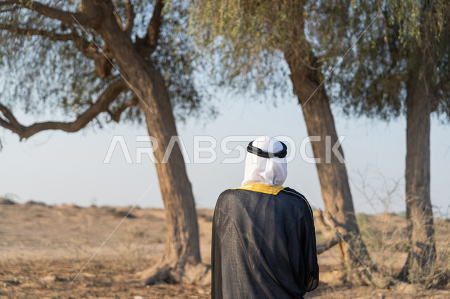 الاحتفال بذكرى تأسيس الاتحاد 2 ديسمبر، حب الوطن والاعتزاز به، اليوم الوطني الاماراتي 1971، صورة من الخلف لرجل عربي خليجي إماراتي يرتدي الكندورة والبشت مع الغترة يقف امام شجرة خضراء كبيرة في الصحراء، التجول في المناطق الطبيعية في الامارات العربية المتحدة