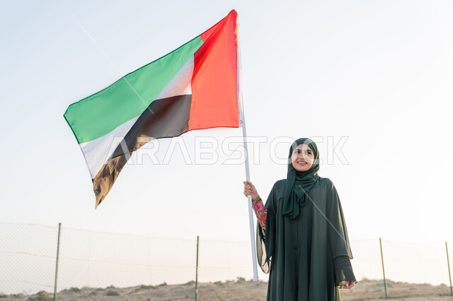 رفع راية الوطن في يوم العلم 3 نوفمبر، استذكار اليوم الوطني الاماراتي 1971، ذكرى تأسيس الاتحاد 2 ديسمبر، إمراة عربية خليجية إماراتية ترتدي الحجاب والعباءة تمسك بيدها علم الإمارات العربية المتحدة تقف في الصحراء في وقت النهار، حب الوطن والاعتزاز به، خلفية الطبيعة الصحراوية