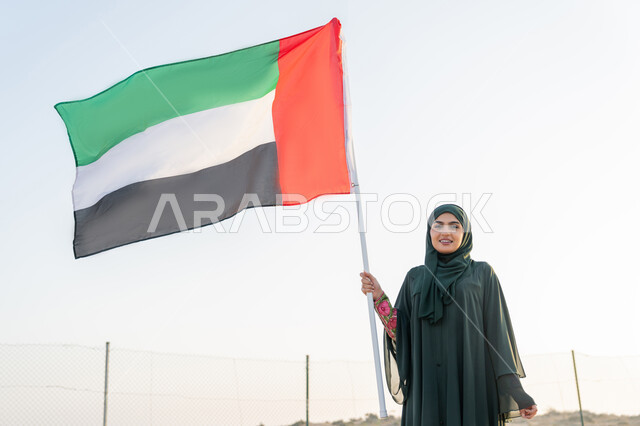 رفع راية الوطن في يوم العلم 3 نوفمبر، استذكار اليوم الوطني الاماراتي 1971، ذكرى تأسيس الاتحاد 2 ديسمبر، إمراة عربية خليجية إماراتية ترتدي الحجاب والعباءة تمسك بيدها علم الإمارات العربية المتحدة تقف في الصحراء في وقت النهار، حب الوطن والاعتزاز به، خلفية الطبيعة الصحراوية