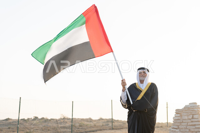 حب الوطن والاعتزاز به، ذكرى تأسيس الاتحاد 2 ديسمبر، رفع راية الوطن في يوم العلم 3 نوفمبر، استذكار اليوم الوطني الاماراتي 1971، رجل عربي خليجي إماراتي يرتدي الكندورة والبشت مع الغترة البيضاء يمسك بيده علم الإمارات العربية المتحدة يقف في منطقة أثرية في الصحراء