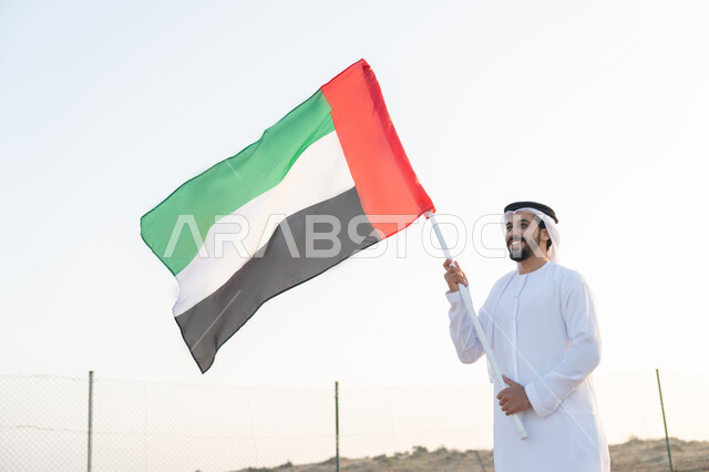 رفع راية الوطن في يوم العلم 3 نوفمبر، استذكار اليوم الوطني الاماراتي 1971، تأسيس الاتحاد 2 ديسمبر، شاب عربي خليجي إماراتي يرتدي الكندورة والغترة يمسك بيده علم الإمارات العربية المتحدة يقف الصحراء في وقت النهار، حب الوطن والاعتزاز به، خلفية الطبيعة الصحراوية