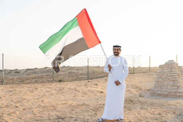 رفع راية الوطن في يوم العلم 3 نوفمبر، استذكار اليوم الوطني الاماراتي 1971، تأسيس الاتحاد 2 ديسمبر، شاب عربي خليجي إماراتي يرتدي الكندورة والغترة يمسك بيده علم الإمارات العربية المتحدة  يقف في منطقة أثرية في الصحراء، حب الوطن والاعتزاز به، خلفية الطبيعة الصحراوية