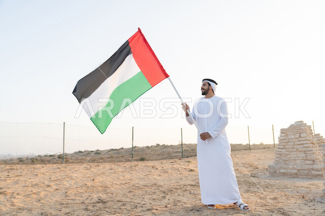 رفع راية الوطن في يوم العلم 3 نوفمبر، استذكار اليوم الوطني الاماراتي 1971، تأسيس الاتحاد 2 ديسمبر، شاب عربي خليجي إماراتي يرتدي الكندورة والغترة يمسك بيده علم الإمارات العربية المتحدة  يقف في منطقة أثرية في الصحراء، حب الوطن والاعتزاز به، خلفية الطبيعة الصحراوية
