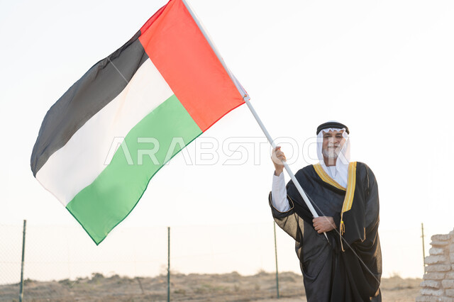 حب الوطن والاعتزاز به، ذكرى تأسيس الاتحاد 2 ديسمبر، رفع راية الوطن في يوم العلم 3 نوفمبر، استذكار اليوم الوطني الاماراتي 1971، رجل عربي خليجي إماراتي يرتدي الكندورة والبشت مع الغترة البيضاء يمسك بيده علم الإمارات العربية المتحدة يقف في منطقة أثرية في الصحراء