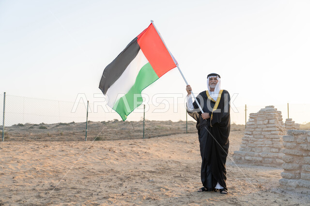 ذكرى تأسيس الاتحاد 2 ديسمبر، رفع راية الوطن في يوم العلم 3 نوفمبر، استذكار اليوم الوطني الاماراتي 1971، رجل عربي خليجي إماراتي يرتدي الكندورة والبشت مع الغترة البيضاء يمسك بيده علم الإمارات العربية المتحدة  يقف في منطقة أثرية في الصحراء، حب الوطن والاعتزاز به