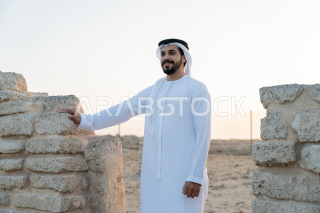 Visiting ancient historical places, remembering the UAE National Day 1971, the founding of the union on December 2, a young Emirati Gulf Arab man wearing a white kandura and ghutra standing in an archaeological area in the desert, love of the homeland and belonging to it, expressions and gestures of pride and honor, desert nature background