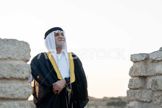 تعابير الفخر والاعتزاز، التجول في الاماكن التاريخية القديمة، اليوم الوطني الاماراتي 1971، ذكرى تأسيس الاتحاد 2 ديسمبر، صورة مقربة لرجل عربي خليجي إماراتي يرتدي الكندورة والبشت مع الغترة يقف في منطقة أثرية في الصحراء، حب الوطن والانتماء اليه، خلفية الطبيعة