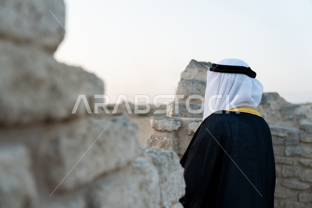 Visiting ancient historical places, UAE National Day 1971, celebrating the anniversary of the founding of the union on December 2, a picture from the back of an Emirati Gulf Arab man wearing a kandura and a bisht with a ghutra standing in an archaeological area in the desert, love of the homeland and belonging to it, expressions of pride and honor, nature background