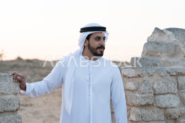 Visiting ancient historical places, remembering the UAE National Day 1971, the founding of the union on December 2, a young Emirati Gulf Arab man wearing a white kandura and ghutra standing in an archaeological area in the desert, love of the homeland and belonging to it, expressions and gestures of pride and honor, desert nature background