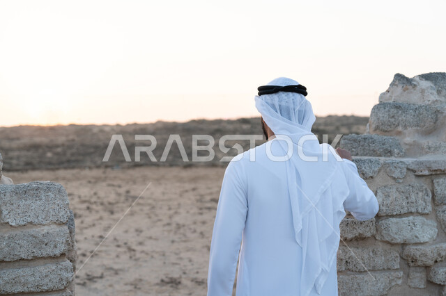 UAE National Day 1971, Visiting ancient historical places, Celebrating the anniversary of the founding of the Union on December 2, Back view of a young Emirati Gulf Arab man wearing a kandura and ghutra standing in an archaeological area in the desert, love of the homeland and belonging to it, desert nature background