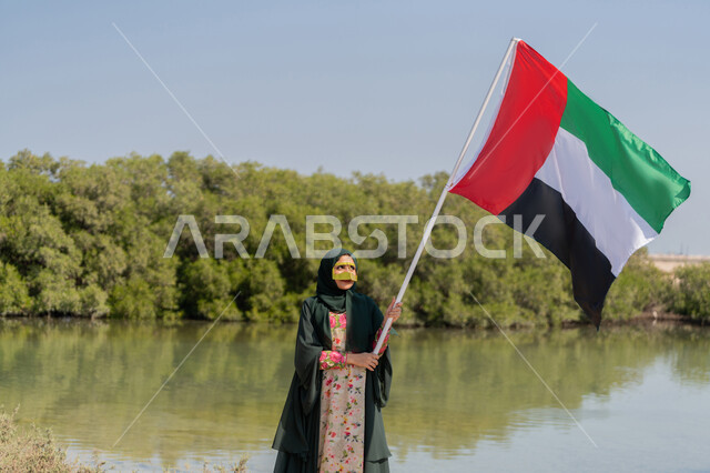استذكار اليوم الوطني الاماراتي 1971، ذكرى تأسيس الاتحاد  2 ديسمبر، إمراة عربية خليجية إماراتية ترتدي الحجاب والعباءة والبرقع الذهبي تمسك بيدها علم الإمارات العربية المتحدة تقف امام بركة المياه والاشجار الخضراء، يوم العلم 3 نوفمبر، حب الوطن والاعتزاز به، خلفية الطبيعة