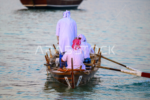 نشاط ترفيهي ممتع, ممارسة هواية الصيد, الاستمتاع بالرحلة البحرية، صورة من الخلف لرجال عرب خليجيون قطريون يرتدون الثوب التقليدي يبحرون في قارب خشبي على شاطئ مدينة الدوحة، مهرجان كتارا الرابع عشر للمحامل التقليدية, فعاليات وأنشطة تراثية وثقافية