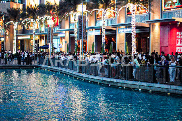 مجموعة من الأشخاص تقف في الساحة الخارجية لدبي مول المطلة على المياه, التصميم الهندسي والديكور المميز على الطراز الحديث للمركز التجاري، مكان ومعلم سياحي مشهور في الإمارات العربية المتحدة, التسوق واستغلال العروض والتخفيض الموسمي، قضاء وقت ممتع داخل المجمعات