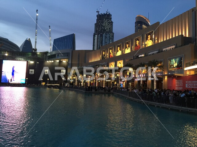Shopping and taking advantage of seasonal offers and discounts, a group of people standing in the outdoor square of Dubai Mall overlooking the water, a famous tourist place and landmark in the United Arab Emirates, the distinctive modern architectural design and decoration of the mall, spending a fun time inside the complexes