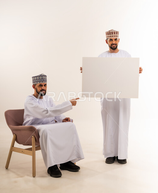 Using illustrations and educational tools, advertisements and marketing offers, blank billboard, portrait of two young Omani Arab twins wearing a dishdasha and a kummah, one of them holding a rectangular mockup and the other pointing at it with his index finger, white background