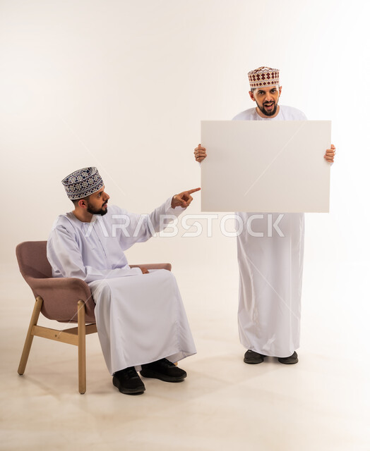 لوح اعلاني فارغ، استخدام لوحات توضيحية ووسائل تعليمية، إعلانات وعروض تسويقية، بورتريه لشابين توأم عربيين خليجيين عمانيين يرتديان الدشداشة والكمة أحدهما يحمل موك اب مستطيل والآخر يشير له بإصبع السبابة، خلفية بيضاء
