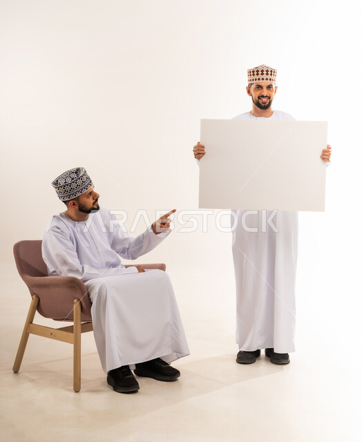 Advertisements and marketing offers, using explanatory panels and educational tools, blank billboard, portrait of two young Omani Arab twins wearing a dishdasha and a kummah, one of them holding a rectangular mockup and the other pointing at it with his index finger, white background