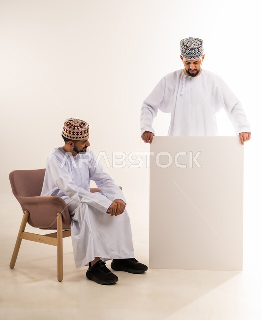 لوح اعلاني فارغ، استخدام لوحات توضيحية ووسائل تعليمية، إعلانات وعروض تسويقية، بورتريه لشابين توأم عربيين خليجيين عمانيين يرتديان الدشداشة والكمة أحدهما يحمل موك اب مستطيل والآخر يجلس على الكرسي، خلفية بيضاء
