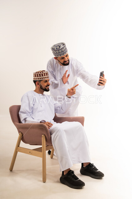 تعابير إلقاء السلام والتحية, استخدام جهاز تقني حديث ومتطور, التواصل مع الأصدقاء والعائلة, بورتريه لشابين عربيين خليجيين عمانيين يرتديان الدشداشة والكمة يقومان بإجراء مكالمة فيديو عبر الهاتف المحمول, خلفية بيضاء