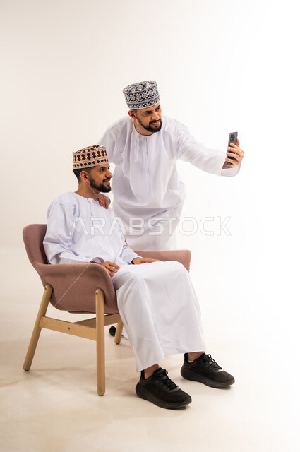 توثيق اللحظات السعيدة عبر الهاتف المحمول, استخدام جهاز تقني حديث ومتطور, قضاء الوقت الممتع مع الاخوة, بورتريه لشاب عربي خليجي عماني يرتدي الدشداشة والكمة يقوم بالتقاط صور السيلفي التذكارية مع أخيه التوأم عبر الجوال, خلفية بيضاء