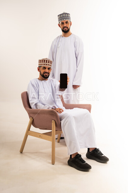 Using a modern and advanced technical device, entertainment and recreation in free time, browsing social media, portrait of a young Arab Gulf Omani man wearing a dishdasha and a kummah sitting in front of his twin brother and holding a mobile phone in his hand, displaying a blank black screen via mobile, white background