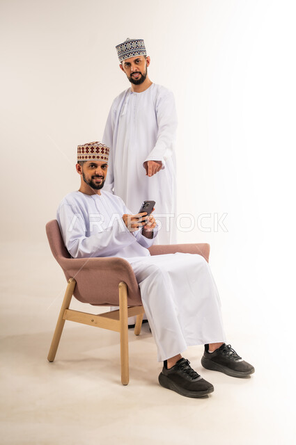 التسلية والترفيه في وقت الفراغ, استخدام جهاز تقني حديث ومتطور, تصفح وسائل التواصل الاجتماعي, بورتريه لشابين توأم عربيين خليجيين عمانيين يرتديان الدشداشة والكمة أحدهما يستعمل الهاتف المحمول والآخر يشير إليه بإصبع السبابة, خلفية بيضاء