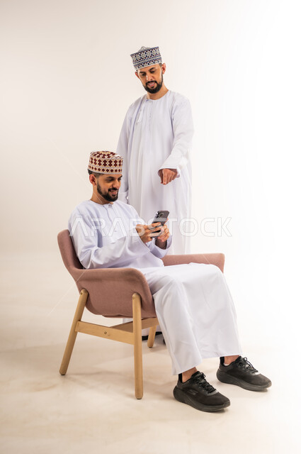 استخدام جهاز تقني حديث ومتطور, التسلية والترفيه في وقت الفراغ, تصفح وسائل التواصل الاجتماعي, بورتريه لشابين توأم عربيين خليجيين عمانيين يرتديان الدشداشة والكمة أحدهما يستعمل الهاتف المحمول والآخر يشير إليه بإصبع السبابة, خلفية بيضاء