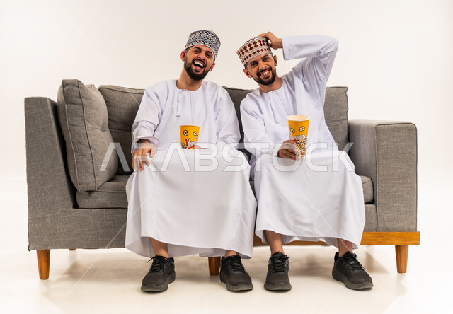 الاستمتاع بمشاهدة الأفلام, تعابير السعادة والضحك الشديد, قضاء الوقت المسلي والممتع مع الأخوة, بورتريه لشابين توأم عربيين خليجيين عمانيين يرتديان الدشداشة والكمة يجلسان على الأريكة ويمسكان علب الفوشار, خلفية بيضاء