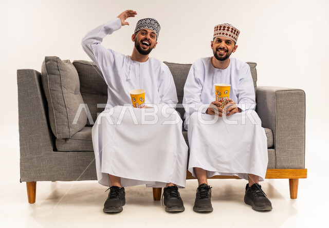 تعابير السعادة والضحك الشديد, الاستمتاع بمشاهدة الأفلام, قضاء الوقت المسلي والممتع مع الأخوة, بورتريه لشابين توأم عربيين خليجيين عمانيين يرتديان الدشداشة والكمة يجلسان على الأريكة ويمسكان علب الفوشار, خلفية بيضاء