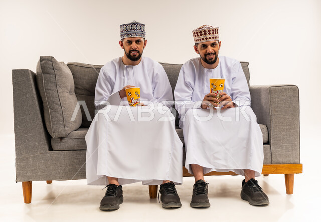 قضاء الوقت المسلي والممتع مع الأخوة, الاستمتاع بمشاهدة الأفلام, تعابير الاندماج والتركيز, بورتريه لشابين توأم عربيين خليجيين عمانيين يرتديان الدشداشة والكمة يجلسان على الأريكة ويمسكان علب الفوشار, خلفية بيضاء