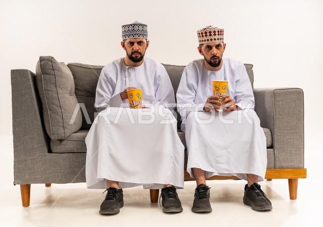 قضاء الوقت المسلي والممتع مع الأخوة, الاستمتاع بمشاهدة الأفلام, تعابير الاندماج والتركيز, بورتريه لشابين توأم عربيين خليجيين عمانيين يرتديان الدشداشة والكمة يجلسان على الأريكة ويمسكان علب الفوشار, خلفية بيضاء