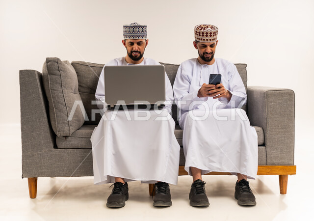 تصفح وسائل التواصل الاجتماعي, استخدام الأجهزة التقنية الحديثة والمتطورة, إنجاز مهام العمل عن بعد, بورتريه لشابين توأم عربيين خليجيين عمانيين يرتديان الدشداشة والكمة يجلسان على الأريكة ويستعملان جهازي الحاسوب المحمول والجوال, خلفية بيضاء