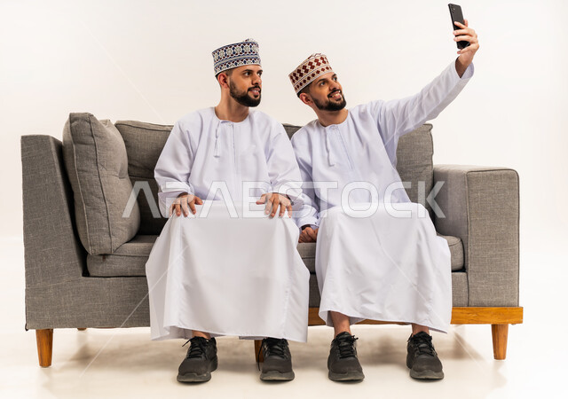 توثيق اللحظات السعيدة عبر الهاتف المحمول, استخدام جهاز تقني حديث ومتطور, قضاء الوقت الممتع مع الاخوة, بورتريه لشاب عربي خليجي عماني يرتدي الدشداشة والكمة يجلس على الأريكة مع أخيه التوأم ويقوم بالتقاط صور السيلفي التذكارية عبر الجوال, خلفية بيضاء