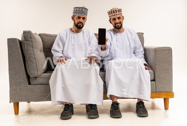 النظر إلى الكاميرا بإيماءات السعادة والسرور, استخدام جهاز تقني حديث ومتطور, قضاء الوقت الممتع مع الأخوة, بورتريه لشاب عربي خليجي عماني يرتدي الدشداشة والكمة يجلس على الأريكة مع أخيه التوأم ويمسك الهاتف المحمول, عرض شاشة سوداء فارغة عبر الجوال, خلفية بيضاء