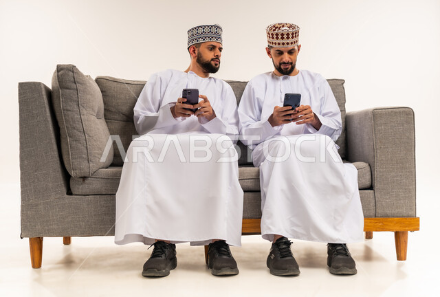 التطفل والنظر خلسة للجوال الآخر, استخدام جهاز تقني حديث ومتطور, تصفح وسائل التواصل الاجتماعي, بورتريه لشابين توأم عربيين خليجيين عمانيين يرتديان الدشداشة والكمة يجلسان على الأريكة ويقومان باستعمال الهاتف المحمول, خلفية بيضاء