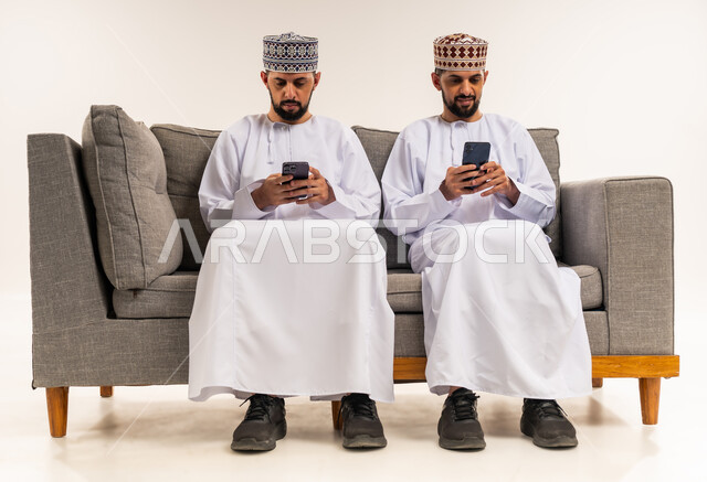 Browsing social media, using a modern and advanced technical device, entertainment and leisure time, portrait of two young Omani Gulf Arab twins wearing a dishdasha and a thobe sitting on a couch and using a mobile phone, white background