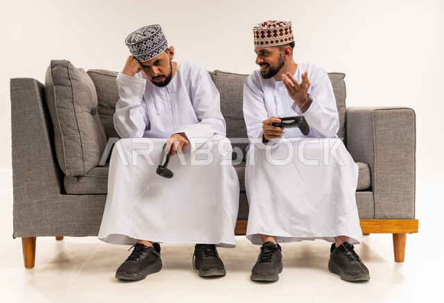 تعابير الحزن لخسارة اللعبة, استخدام التكنولوجيا الحديثة للتسلية والترفيه, الاستمتاع بألعاب الفيديو الممتعة, الفرح للفوز والنصر, بورتريه لشابين توأم عربيين خليجيين عمانيين يرتديان الدشداشة والكمة يجلسان على الأريكة ويمسكان بأيديهم عصا التحكم, خلفية بيضاء