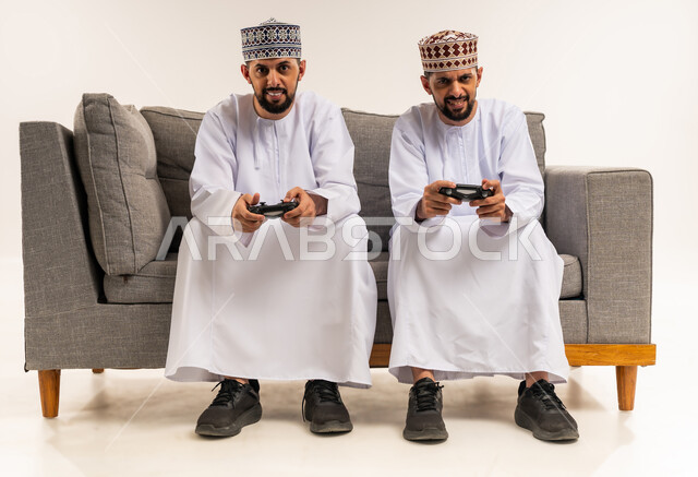 قضاء الوقت الممتع مع الأخوة, استخدام التكنولوجيا الحديثة للتسلية والترفيه, الاستمتاع بألعاب الفيديو الممتعة, بورتريه لشابين توأم عربيين خليجيين عمانيين يرتديان الدشداشة والكمة يجلسان على الأريكة ويمسكان بأيديهم عصا التحكم, خلفية بيضاء