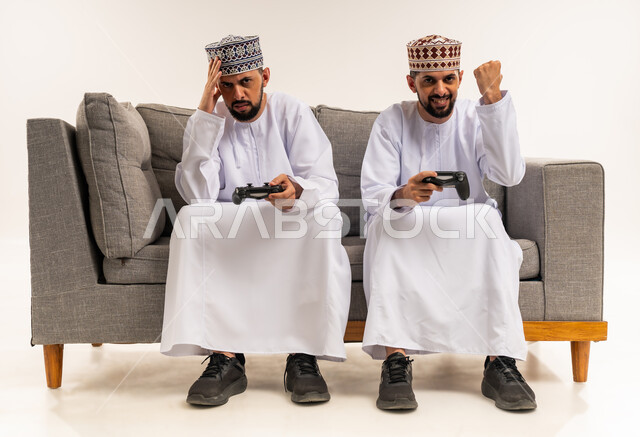 الفرح للفوز والنصر, تعابير الحزن لخسارة اللعبة, استخدام التكنولوجيا الحديثة للتسلية والترفيه, الاستمتاع بألعاب الفيديو الممتعة, بورتريه لشابين توأم عربيين خليجيين عمانيين يرتديان الدشداشة والكمة يجلسان على الأريكة ويمسكان بيديهم عصا التحكم, خلفية بيضاء