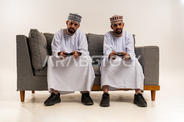 Using modern technology for entertainment and fun, spending quality time with brothers, enjoying fun video games, portrait of two young Omani Gulf Arab twins wearing a dishdasha and a thobe sitting on a couch holding a joystick in their hands, white background