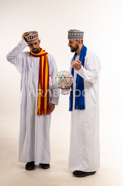 تعابير الفرح بالنصر والفوز، إيماءات الحزن لخسارة الفريق، بورتريه لشاب عربي خليجي عماني يرتدي الدشداشة والكمة يقوم بإغاظة أخوه التوأم لانتصار فريقه المفضل، ارتداء اوشحة تشجيعية لفرق كرة القدم، المباريات والألعاب الرياضية, الجسم بالطول الكامل, خلفية بيضاء