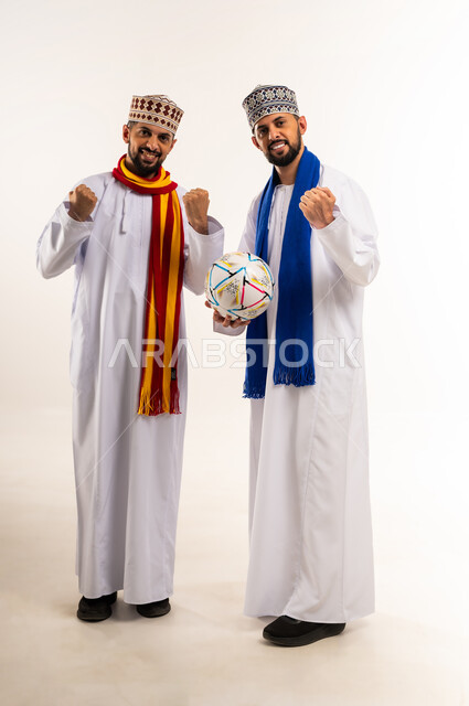 النظر الى الكاميرا بإيماءات الحماس و الشغف، المباراة والألعاب الرياضية، الفرح لانتصار الفريق، بورتريه لشابين توأم عربيين خليجيين عمانيين يرتديان الدشداشة والكمة مع أوشحة تشجيعية لفرق كرة القدم يرفعان قبضة يديهما بتعابير الفوز, الجسم بالطول الكامل، خلفية بيضاء