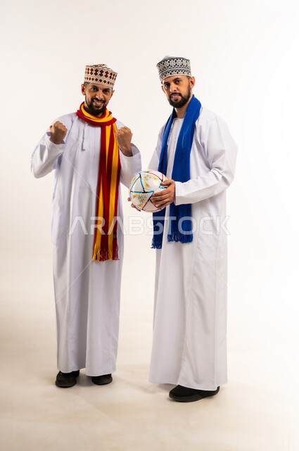 المباراة والألعاب الرياضية, النظر الى الكاميرا بإيماءات الحماس والتشجيع، الفرح لانتصار الفريق، بورتريه لشابين توأم عربيين خليجيين عمانيين يرتديان الدشداشة والكمة مع أوشحة تشجيعية لفرق كرة القدم يرفعان قبضة يديهما بتعابير الفوز, الجسم بالطول الكامل، خلفية بيضاء