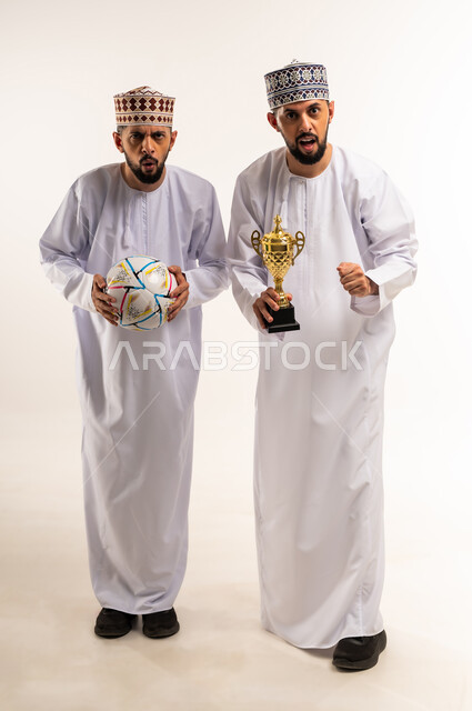 النظر الى الكاميرا بإيماءات الحماس و التشجيع, الفرح لانتصار الفريق المفضل، المباريات والألعاب الرياضية، بورتريه لشابين توأم عربيين خليجيين عمانيين يرتديان الدشداشة والكمة يحملان بأيديهم الكأس وكرة القدم, الجسم بالطول الكامل، خلفية بيضاء
