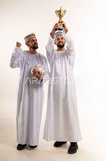 رفع قبضة اليد بإيماءات الحماس و التشجيع, الفرح لانتصار الفريق المفضل، المباراة والألعاب الرياضية، بورتريه لشابين توأم عربيين خليجيين عمانيين يرتديان الدشداشة والكمة يحملان بأيديهم الكأس وكرة القدم, الجسم بالطول الكامل، خلفية بيضاء
