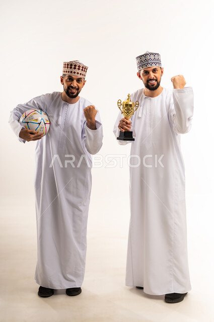 رفع قبضة اليد بتعابير الفرح لانتصار الفريق المفضل, النظر الى الكاميرا بإيماءات الحماس و الشغف، المباراة والألعاب الرياضية، بورتريه لشابين توأم عربيين خليجيين عمانيين يرتديان الدشداشة والكمة يحملان بأيديهم الكأس وكرة القدم, الجسم بالطول الكامل، خلفية بيضاء